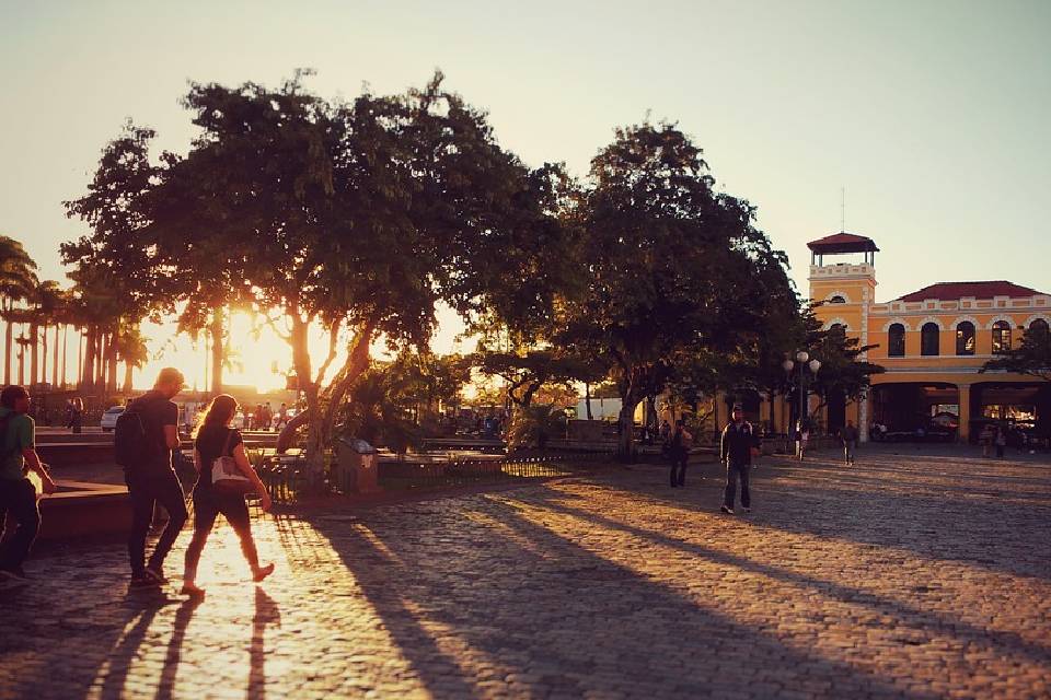 Foto colorida com pessoas caminhando e o Sol se pondo por trás das árvores