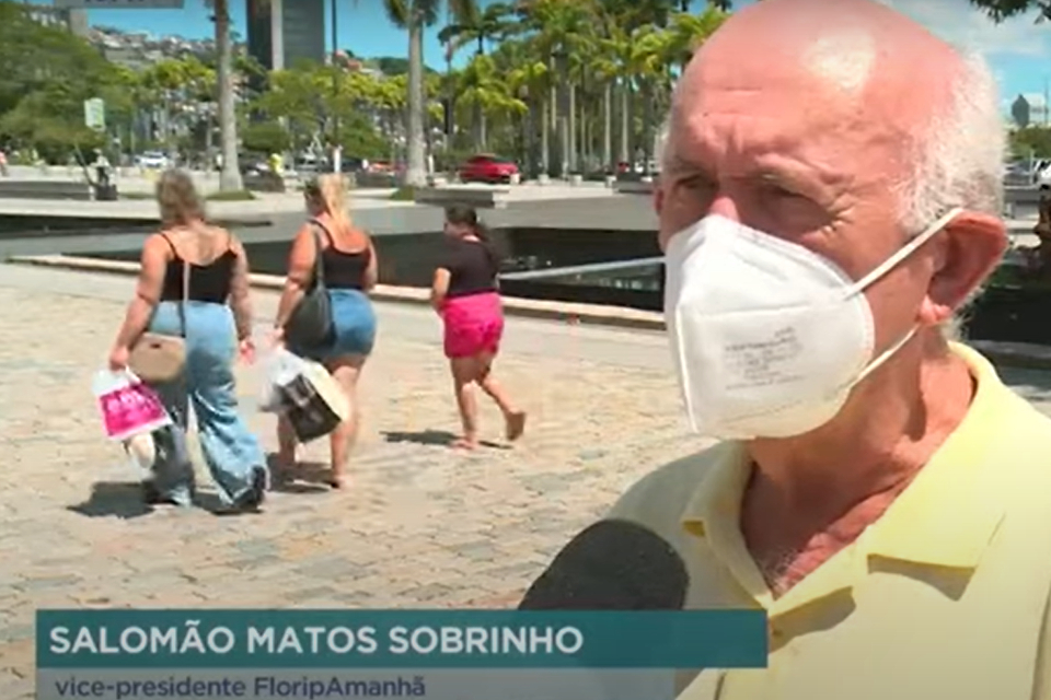 Foto colorida com palmeiras ao fundo e um homem calvo de cabelos grisalhos sendo entrevistado