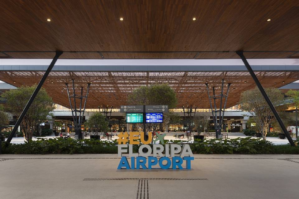 Foto colorida com fundo em estruturas de madeira marrom e um letreiro à frente com a inscrição "#eufloripaairport"