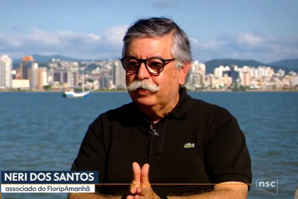 Foro colorida de um homem branco de meia idade com cabelos grisalhos, bigode grisalho e camiseta preta e ao fundo o mar s