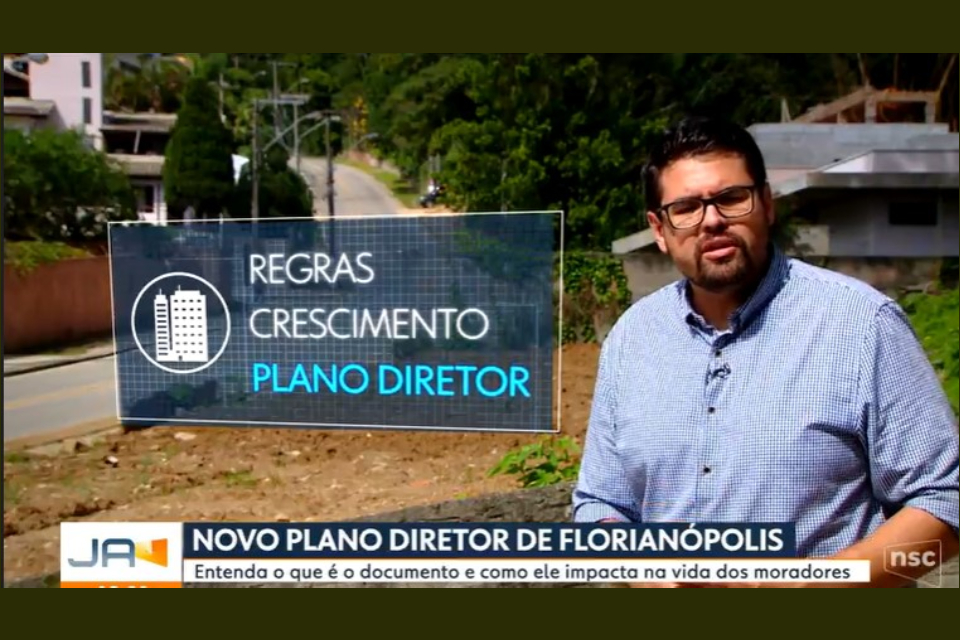 Foto colorida de um terreno urbano com um repórter de camisa azul clara de óculos, cabelos e barba preta