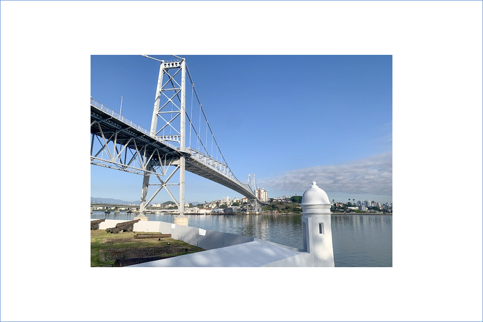 Foto colorida de uma torre de um forte antigo com uma ponte de ferro e o mar ao fundo