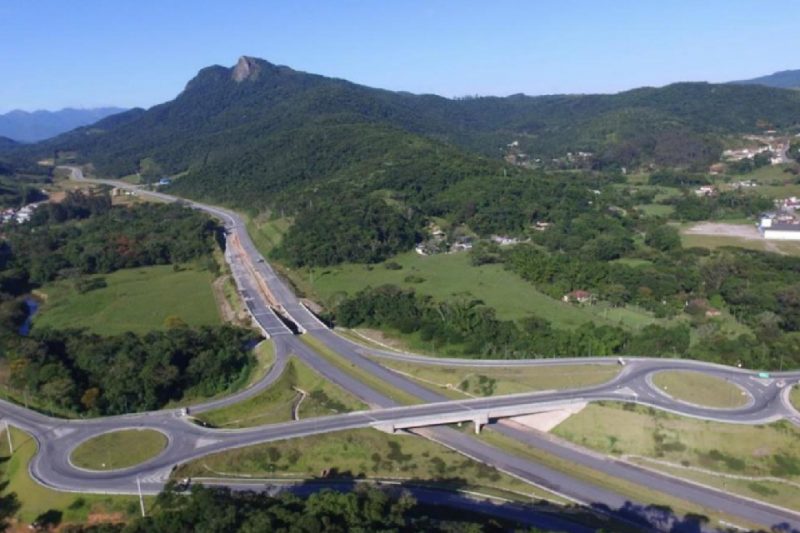 Foto colorida com vista aérea de uma paisagem com morro coberto por vegetação, rodovia com um viaduto e duas rótulas do contorno viário