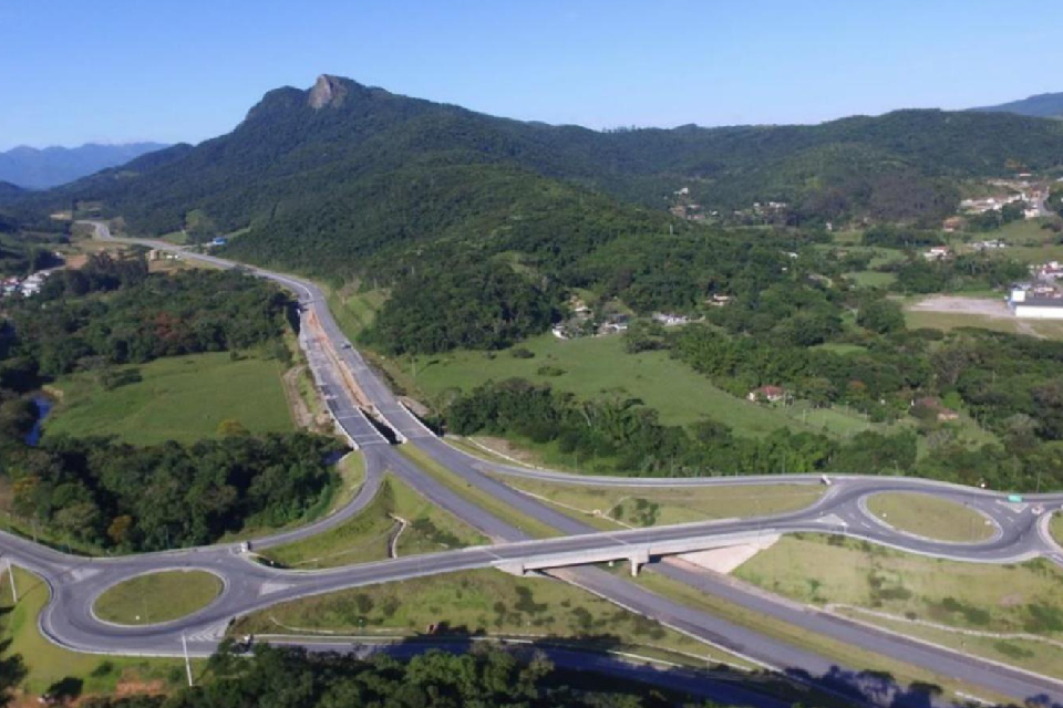 Foto colorida com vista aérea de uma paisagem com morro coberto por vegetação, rodovia com um viaduto e duas rótulas do contorno viário