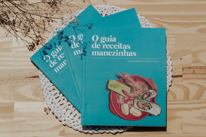 Na imagem um livro de receitas culinárias com a capa na cor azul claro e um prato com frutos do mar