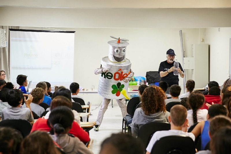 Foto colorida em uma sala de aula com várias crianças e um robô de lata prateado