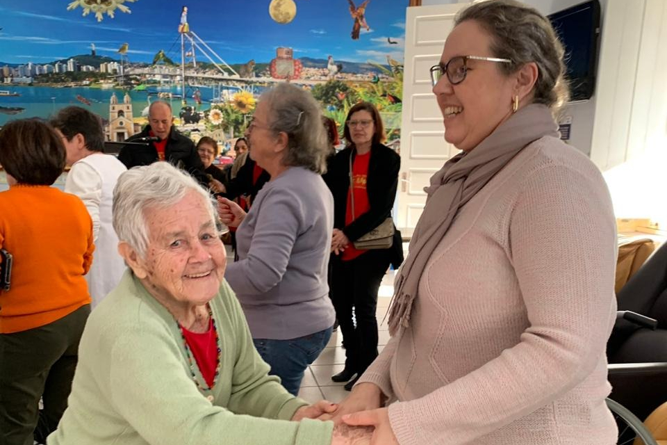 Foto colorida de uma senhora idosa dançando com uma outra mulher mais nova em um asilo