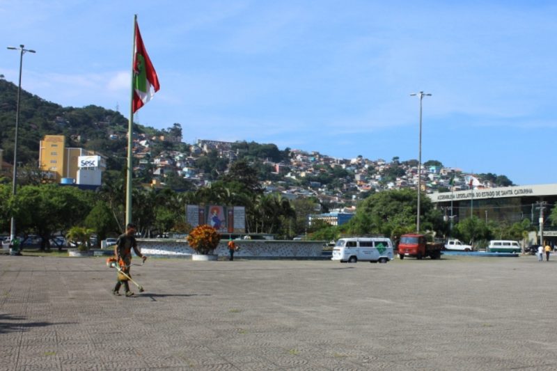 Foto colorida de uma praça com piso de concreto, um trabalhador fazendo manutenção com a bandeira de Santa Catarina ao fundo