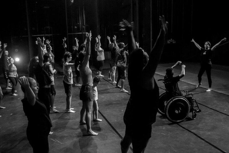 Imagem em preto e branco com várias pessoas com as mãos para cima fazendo uma evolução de dança