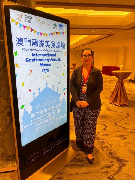 A imagem mostra uma mulher de óculos, sorridente, usando um blazer preto e uma calça estampada, posando ao lado de um totem eletrônico. O totem exibe informações sobre o "Fórum Internacional de Gastronomia, Macau," que ocorreu em 17 de junho. O texto no totem está escrito em chinês, português e inglês. O fundo do local tem um ambiente bem iluminado, com mesas altas cobertas por toalhas e decoração sofisticada, sugerindo que o evento é realizado em um local elegante e apropriado para conferências.