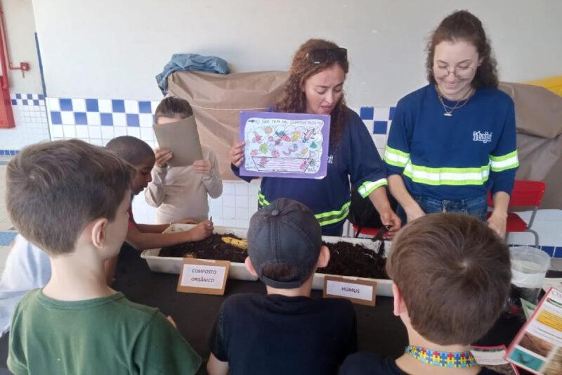 Imagem de uma atividade de educação ambiental com crianças. Duas mulheres, usando uniformes da CASAN, estão em uma mesa realizando uma oficina sobre compostagem. Uma delas segura um cartaz colorido que diz "O que tem na compostagem" e mostra ilustrações de itens compostáveis. A outra está manipulando o composto orgânico na mesa. Ao redor da mesa, há várias crianças observando e participando. Na mesa, há recipientes rotulados como "Composto Orgânico" e "Húmus". As crianças parecem interessadas e envolvidas na atividade.