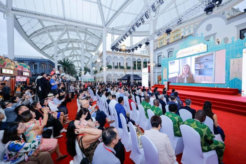 A imagem mostra um grande evento realizado em um local coberto e arejado, com uma plateia de várias pessoas sentadas em cadeiras brancas voltadas para um palco. No palco, há um grande telão exibindo uma mulher falando em uma videoconferência. O ambiente é iluminado e decorado com elementos arquitetônicos ao fundo, incluindo uma construção de estilo clássico. À esquerda, há uma área com estandes de comida e várias pessoas em pé, algumas tirando fotos e outras observando. O evento parece ser formal, com muitos participantes vestindo trajes elegantes.