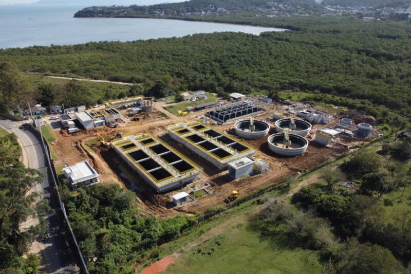 A imagem mostra uma vista aérea de um complexo de construção, possivelmente uma estação de tratamento de esgoto, localizada próxima a uma área costeira com vista para o mar. O complexo é composto por várias estruturas grandes, incluindo tanques circulares e retangulares. A área ao redor é predominantemente verde com vegetação densa. No canto esquerdo, há uma estrada que passa ao lado do complexo. Ao fundo, há uma área de floresta densa e algumas colinas, com uma cidade visível ao longe. A construção parece estar em andamento, com máquinas e equipamentos espalhados pelo local.