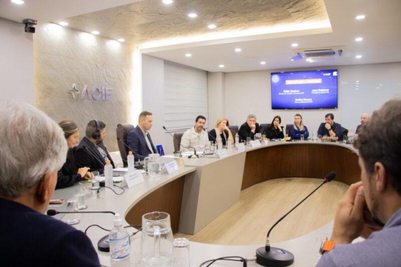 A imagem mostra uma reunião formal na sede da ACIF (Associação Comercial e Industrial de Florianópolis). As pessoas estão sentadas ao redor de uma mesa de conferência em formato de U, equipada com microfones, copos de água e materiais de anotação. No centro da mesa, um homem em destaque está falando ao microfone. Ao fundo, uma tela exibe informações sobre a pauta da reunião, incluindo os nomes dos participantes: Fábio Queiroz, Jaime Batistot, e Bedida Ramos. A sala é bem iluminada com luzes embutidas no teto e um painel decorativo na parede com o logotipo da ACIF à esquerda. As pessoas presentes parecem estar atentas e envolvidas na discussão, refletindo um ambiente de trabalho colaborativo e profissional.