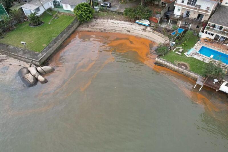 Vista aérea de uma pequena praia cercada por casas e áreas verdes, com a água manchada de laranja devido à presença de algas, destacando a intervenção humana e os efeitos no ambiente costeiro.