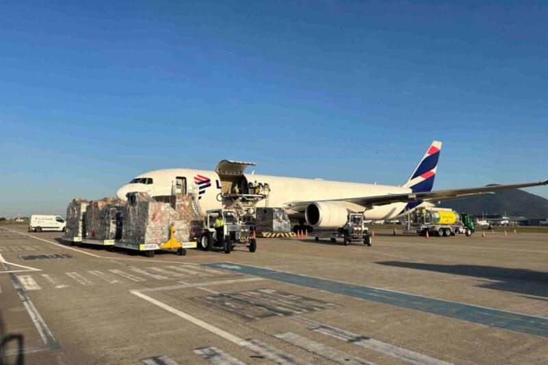 A imagem mostra um avião de carga estacionado em uma pista de aeroporto sob um céu claro e azul. O avião, com a porta de carga aberta, está sendo carregado com grandes paletes de mercadorias cobertas por lonas protetoras. Veículos de suporte e carregamento estão posicionados ao lado do avião, ajudando na movimentação das mercadorias. No fundo, há outros veículos e partes da infraestrutura do aeroporto visíveis. Uma montanha é vista ao fundo, adicionando ao cenário.