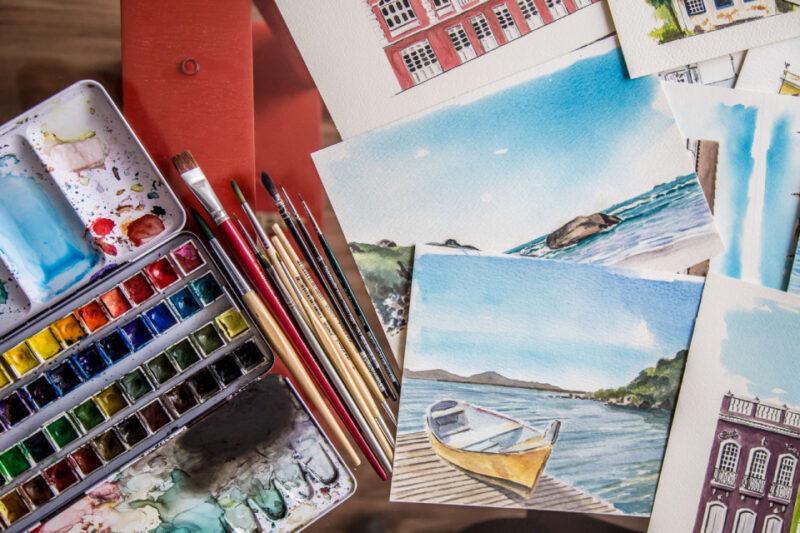 Vista de cima de uma mesa de trabalho de arte com várias aquarelas, pincéis e uma paleta de tintas. As pinturas incluem paisagens costeiras com barcos e cenas urbanas com prédios históricos. A paleta de tintas mostra diversas cores usadas e os pincéis estão organizados ao lado das pinturas.