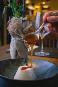 "Uma mão segurando uma colher derrama calda de caramelo sobre um pudim branco em um prato preto. Ao fundo, um vaso com flores embrulhadas em tecido rústico adiciona um toque decorativo à mesa de jantar."