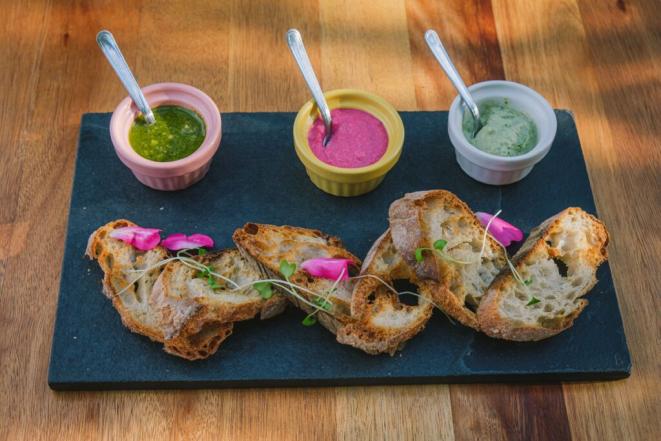 "Uma tábua de ardósia com fatias de pão rústico acompanhadas de três potes de molho coloridos: pesto verde, beterraba rosa e um creme de ervas claro. As fatias de pão são decoradas com flores comestíveis, criando uma apresentação atraente."