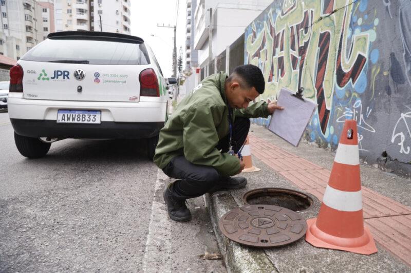 Homem vestido com jaqueta verde e calça preta, agachado ao lado de um bueiro aberto na calçada, segurando uma prancheta com papéis. Ao lado dele, há dois cones de sinalização laranja e branco. Atrás do homem, um carro branco com a logo 'JPR Ambiental' e um mural com grafites coloridos na parede. A rua é ladeada por edifícios altos.