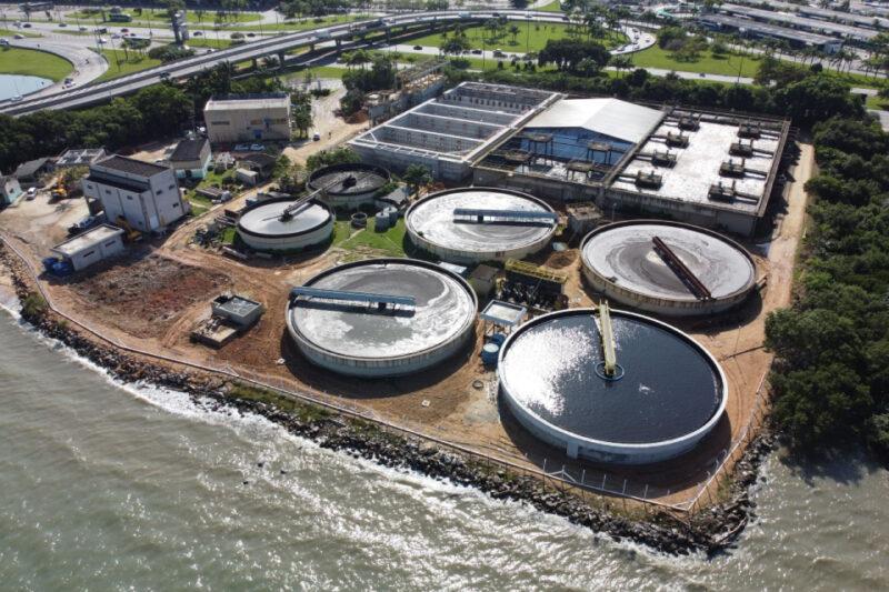 "Vista aérea de uma estação de tratamento de água localizada à beira-mar, com vários tanques circulares grandes e edificações menores ao redor. A estação está cercada por vegetação e próxima a rodovias e áreas urbanas ao fundo."