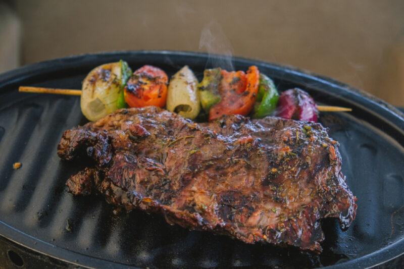 "Uma suculenta peça de carne grelhada está sendo preparada em uma grelha ao lado de um espeto de vegetais coloridos, incluindo cebolas, tomates e pimentões. A carne e os vegetais estão ligeiramente queimados, indicando que estão quase prontos para servir."