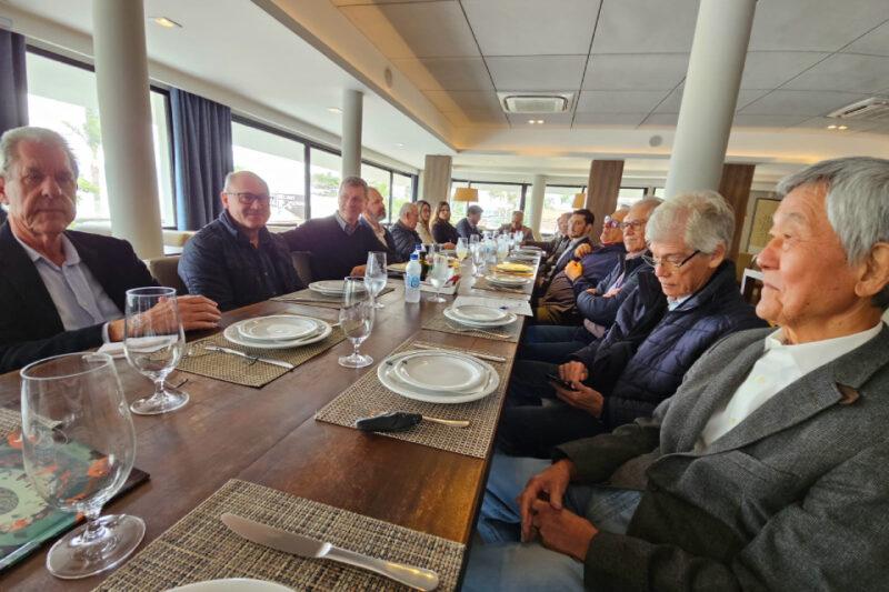 A imagem mostra uma reunião formal com várias pessoas sentadas ao redor de uma longa mesa de jantar. À esquerda, um homem está falando ao microfone, segurando um papel, enquanto os demais participantes escutam atentamente. Na mesa, há pratos, copos e talheres organizados, sugerindo que a reunião está ocorrendo antes de uma refeição. As janelas amplas permitem a entrada de luz natural, iluminando a sala. A atmosfera é séria e profissional, com os participantes focados na discussão.