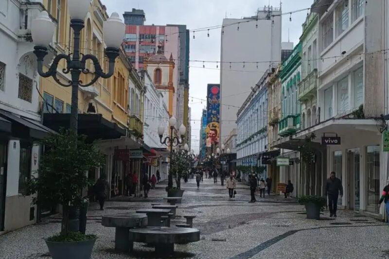 Rua de pedestres com calçada de mosaico em preto e branco, cercada por edifícios históricos coloridos e luminárias clássicas, com pessoas caminhando e um mural colorido ao fundo.