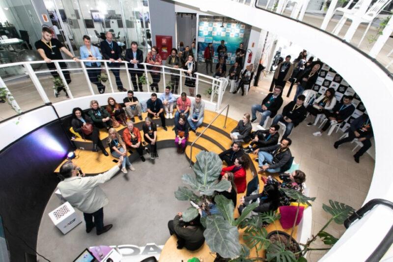 "Imagem de um evento em um espaço moderno e bem iluminado, com pessoas sentadas em um auditório circular. Um palestrante, visto de costas, fala para o público. As pessoas estão atentas e há algumas plantas ao redor do ambiente, que possui decoração clean e elementos tecnológicos."