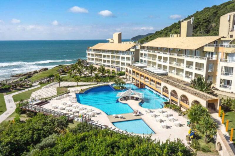 Vista aérea de um resort à beira-mar com uma grande piscina em formato irregular no centro, cercada por espreguiçadeiras e mesas com guarda-sóis brancos. O resort possui várias alas de edifícios de três andares com varandas voltadas para a piscina e o mar. Ao fundo, vê-se a praia com ondas quebrando suavemente e uma área arborizada com colinas verdejantes. O céu está claro com algumas nuvens, indicando um dia ensolarado.