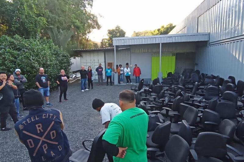 Foto de um grupo de pessoas reunidas em um pátio ao ar livre, ao lado de uma construção de estrutura metálica cinza. À direita da imagem, há muitas cadeiras de escritório pretas empilhadas. As pessoas estão em pé em semicírculo, algumas interagindo e outras observando, em um ambiente arborizado ao fundo. A cena ocorre durante o dia, com luz natural.