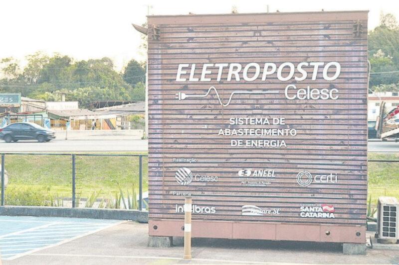 A imagem mostra um eletroposto da Celesc, localizado em Florianópolis. O eletroposto é uma estrutura de madeira com a inscrição "ELETROPOSTO Celesc" e "SISTEMA DE ABASTECIMENTO DE ENERGIA" em letras brancas. Abaixo dessas inscrições, aparecem os logotipos das instituições envolvidas: Celesc, ANEEL, CELETI, Infrabras, WEG, Cooperativa Santa Catarina. À esquerda, há uma estação de recarga de veículos elétricos. Ao fundo, é possível ver uma estrada com alguns veículos em movimento, além de estabelecimentos comerciais e vegetação.