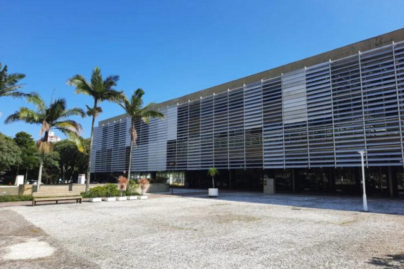 Fachada de um edifício moderno com fachada de vidro e estrutura de brises horizontais em tons de cinza. Em frente ao edifício, há uma praça pavimentada com pedras, palmeiras altas e vasos de plantas. O céu está azul claro, sem nuvens.