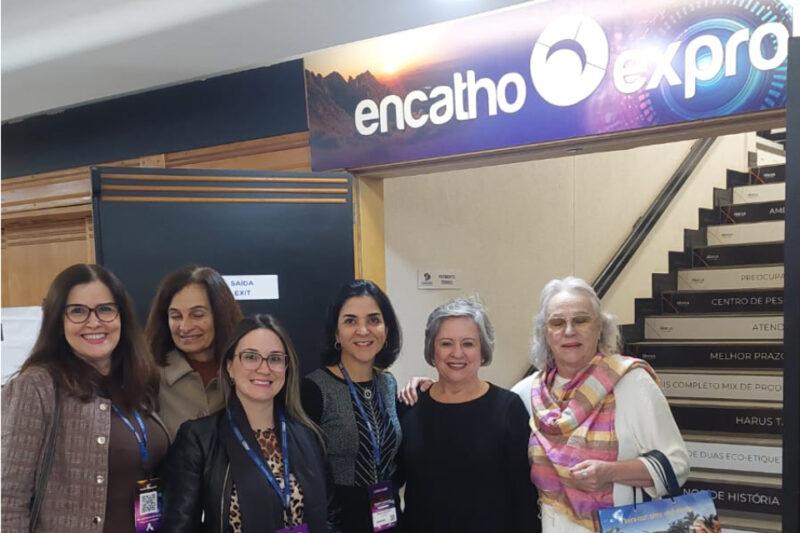 "Seis mulheres posam sorrindo para a foto em frente à entrada do evento Encatho Exprotel. Elas estão de pé sob um banner que exibe o nome do evento, parecendo estar felizes e engajadas. A escada ao lado tem palavras relacionadas à hospitalidade e serviços."