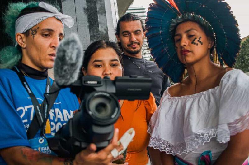 "Quatro pessoas se reúnem ao redor de uma câmera durante um evento. Uma mulher à esquerda, com cabelo verde claro e tatuagens, segura a câmera. Ao lado dela, uma mulher jovem observa, enquanto um homem ao fundo sorri. À direita, uma mulher usa um cocar indígena colorido e tem pintura facial tradicional. Eles parecem concentrados no que estão filmando, destacando a diversidade cultural e o trabalho colaborativo."