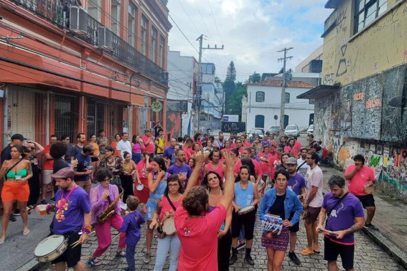 A imagem mostra uma rua movimentada com um grande grupo de pessoas participando de um evento ao ar livre. Muitas delas estão vestidas com camisetas coloridas, predominantemente em tons de rosa e roxo, e algumas estão tocando instrumentos musicais, como tambores e chocalhos. As pessoas parecem estar envolvidas em uma espécie de desfile ou celebração, caminhando pela rua estreita e ladeada por prédios antigos com fachadas coloridas. Há também grafites nas paredes de alguns edifícios. No fundo, pode-se ver outras pessoas assistindo ao evento e algumas placas de trânsito e carros estacionados. A atmosfera é animada e vibrante, com uma mistura de pessoas de todas as idades aproveitando o evento.