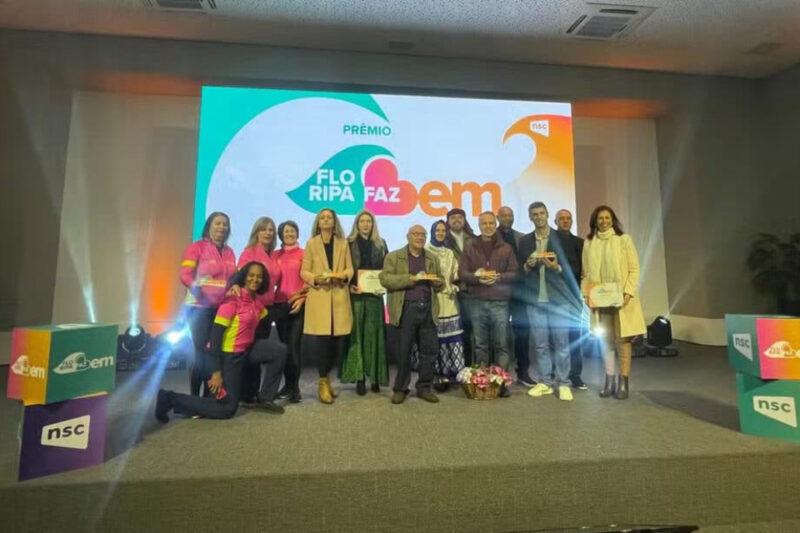 Grupo de pessoas posando para uma foto em um palco durante a premiação 'Floripa Faz Bem'. O grupo inclui mulheres vestindo camisas rosa e calças escuras, e outras pessoas com roupas casuais e formais, segurando troféus e certificados. Ao fundo, há um grande painel com o logotipo do evento 'Floripa Faz Bem'. O palco é iluminado por luzes brancas e coloridas, com cubos decorativos com a marca 'NSC' no canto.