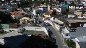 A imagem mostra uma vista aérea de um bairro residencial. Há várias casas e prédios baixos, muitos com telhados de telha cerâmica. Duas ruas principais são visíveis, com ônibus e carros circulando. No centro da imagem, há uma quadra de esportes cercada por grades. A área parece ser densamente povoada, com casas próximas umas das outras e poucas áreas verdes visíveis. A cena parece ocorrer durante o dia, com boa iluminação natural e sombras projetadas pelas construções.