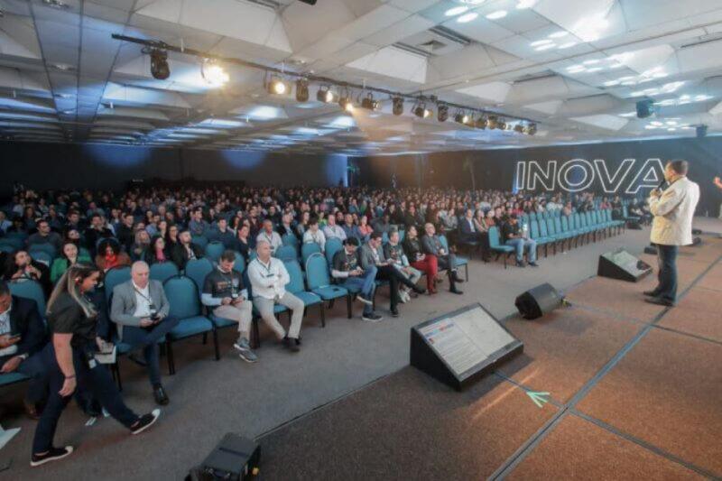 Imagem de uma conferência ou evento corporativo com grande público. Em um auditório bem iluminado, centenas de pessoas estão sentadas em cadeiras azuis, todas voltadas para o palco onde um palestrante, vestido de terno claro, está falando. À direita do palco, há uma grande palavra 'INOVA' escrita na parede. No canto inferior direito, é possível ver uma tela de apresentação e equipamentos de som. A atmosfera é profissional, com iluminação direcionada ao palco.