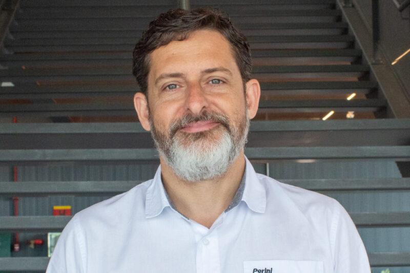 Homem de barba grisalha, cabelo curto e ondulado, veste uma camisa social branca com o logotipo da Perini na altura do peito. Ele está em um ambiente interno com escadas de metal ao fundo, olhando diretamente para a câmera com uma expressão serena.