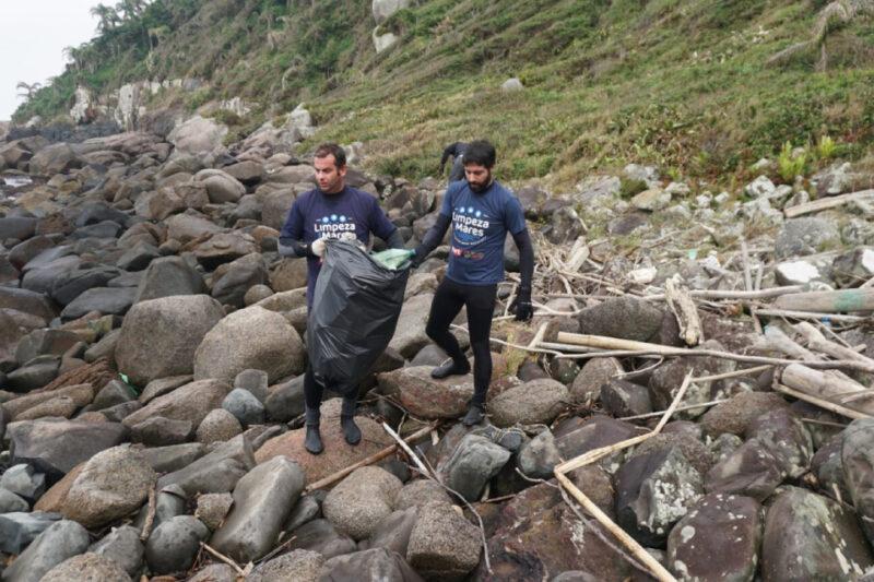 Dois homens participam de uma ação de limpeza em uma área rochosa próxima ao mar. Eles vestem camisetas azuis com o texto "Limpeza dos Mares" e carregam um grande saco de lixo preto. A paisagem ao redor é composta por rochas, galhos espalhados e vegetação rasteira na encosta ao fundo. Ambos usam luvas para proteção enquanto recolhem o lixo.