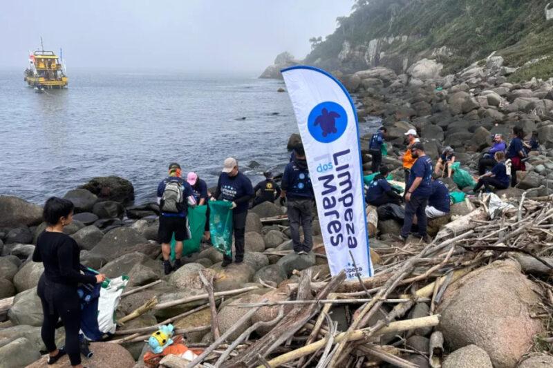 Foto de uma equipe realizando a limpeza de uma área rochosa próxima ao mar. Várias pessoas estão coletando lixo e detritos em sacos verdes. Há uma bandeira vertical com o logotipo do projeto "Limpeza dos Mares" no centro da imagem. Ao fundo, um barco amarelo está ancorado na água. O cenário é composto por pedras e vegetação costeira.