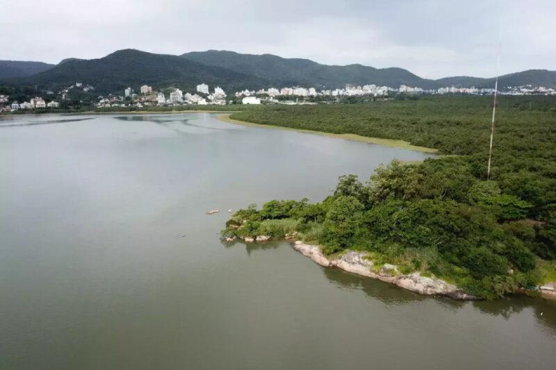 Vista aérea de uma lagoa ou baía cercada por vegetação densa, com uma pequena península arborizada à direita. Ao fundo, uma área urbana com edifícios brancos e uma cadeia de montanhas coberta de vegetação. O céu está nublado.