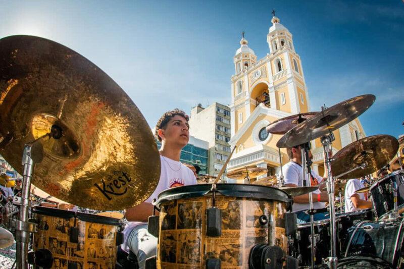 "Um jovem baterista toca em um kit de bateria ao ar livre durante um evento em frente à Catedral Metropolitana de Florianópolis. O céu está claro e a catedral amarela com suas torres gêmeas se destaca ao fundo, criando um cenário vibrante e cultural."