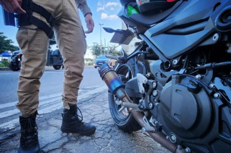 Policial em pé ao lado de uma motocicleta preta, destacando o escapamento, em uma rua com veículos e árvores ao fundo, sob um céu azul.