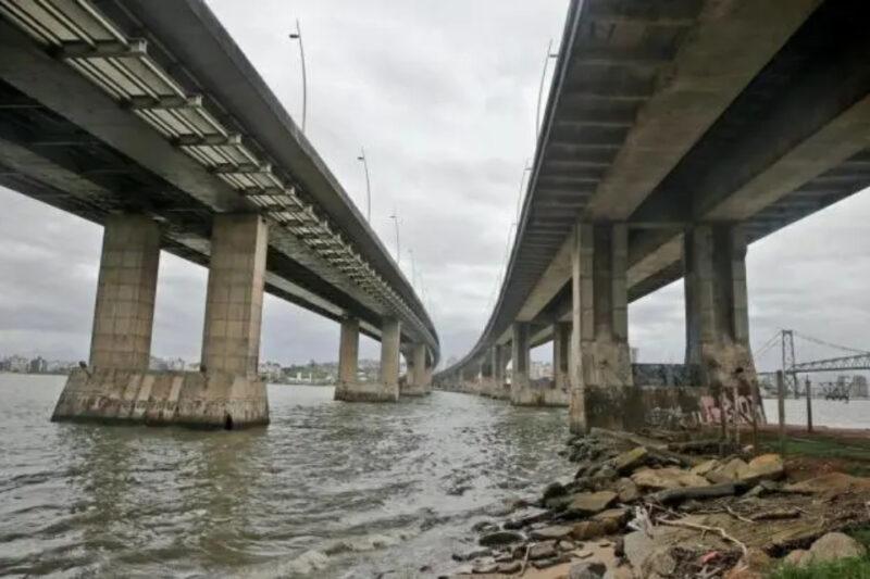 "Foto das pontes de Florianópolis, capturada debaixo das estruturas paralelas que atravessam o mar. A imagem mostra os pilares robustos das pontes sobre a água, com um céu nublado ao fundo. No canto direito da imagem, há uma pequena faixa de terra com pedras e vegetação."