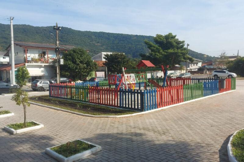 A imagem mostra um parquinho infantil cercado por uma cerca colorida, com brinquedos como escorregador e balanço. O parquinho está situado em uma área pavimentada com pequenos canteiros de plantas ao redor. Ao fundo, há uma rua com carros estacionados e casas residenciais. Além disso, pode-se ver uma área arborizada e montanhas ao longe, indicando que o local está em uma região montanhosa. O dia parece estar ensolarado, com céu claro.