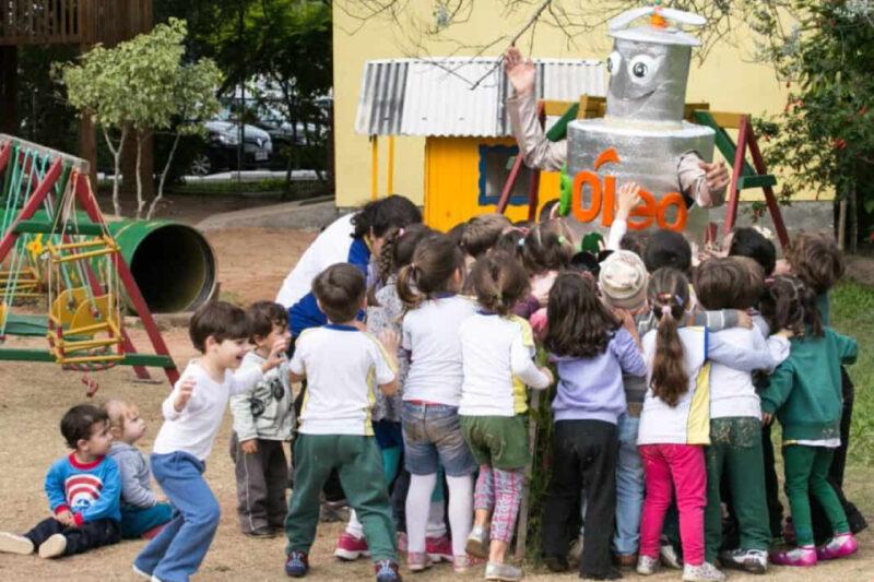 Um grupo de cerca de 20 crianças, vestidas com roupas coloridas e uniformes escolares, rodeia um mascote grande em formato de lata de óleo, que sorri e levanta os braços em um gesto amigável. O mascote tem olhos grandes e um chapéu, e a palavra 'óleo' escrita no peito. As crianças parecem estar abraçando e interagindo com o mascote. A cena acontece em um playground com brinquedos de madeira e um túnel verde ao fundo, com árvores e um edifício amarelo ao redor.