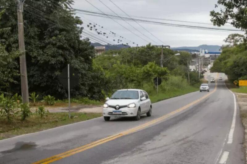 A imagem mostra uma estrada rural asfaltada, com uma faixa amarela contínua no centro e faixas brancas nas bordas. No lado esquerdo, há uma vegetação densa e postes de energia com cabos aéreos. Um carro branco está em destaque, dirigindo na direção da câmera, enquanto outro veículo está mais ao fundo, na mesma faixa. À direita, a estrada é ladeada por mais árvores e uma placa de trânsito amarela. O céu está nublado, sugerindo um dia cinzento. Ao fundo, é possível ver uma área urbana com alguns edifícios e montanhas no horizonte.