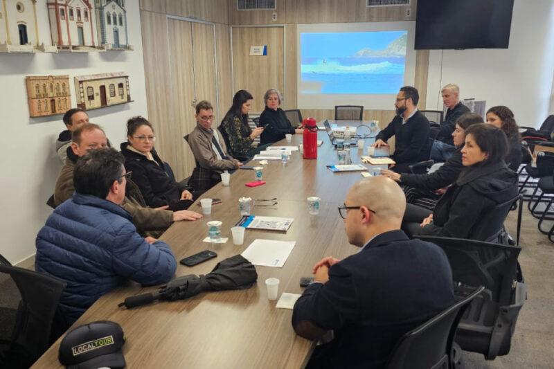 Reunião do Conselho Municipal de Turismo em uma sala de conferências. Cerca de doze pessoas estão sentadas ao redor de uma mesa retangular, discutindo planos estratégicos. Há documentos, copos descartáveis e uma garrafa térmica vermelha sobre a mesa. Na parede, há uma tela de projeção mostrando uma imagem de uma paisagem costeira e quadros com imagens de edifícios históricos.